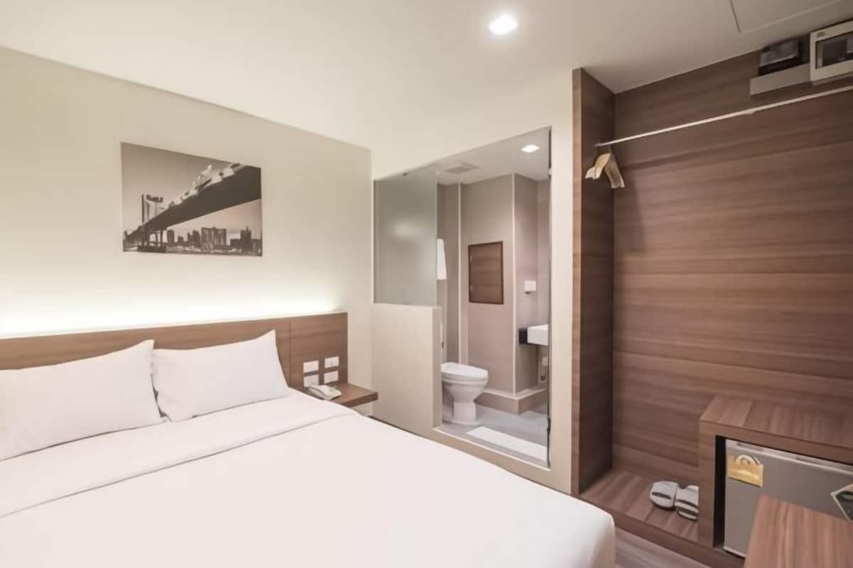 Una habitación de hotel con cama, mesita de noche, espejo, ducha y una pequeña zona de estar.
