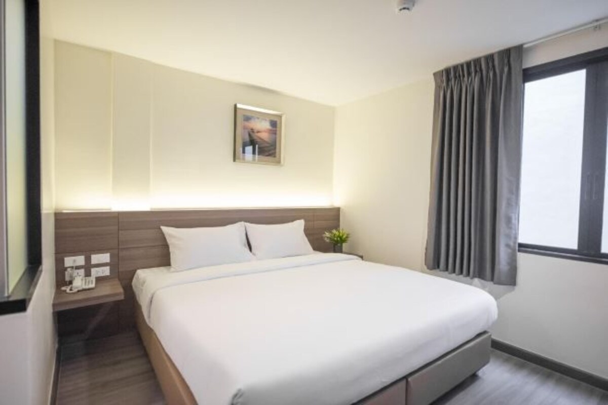 A hotel room with a large bed, a bedside table with a phone, a small plant, and a window with curtains.