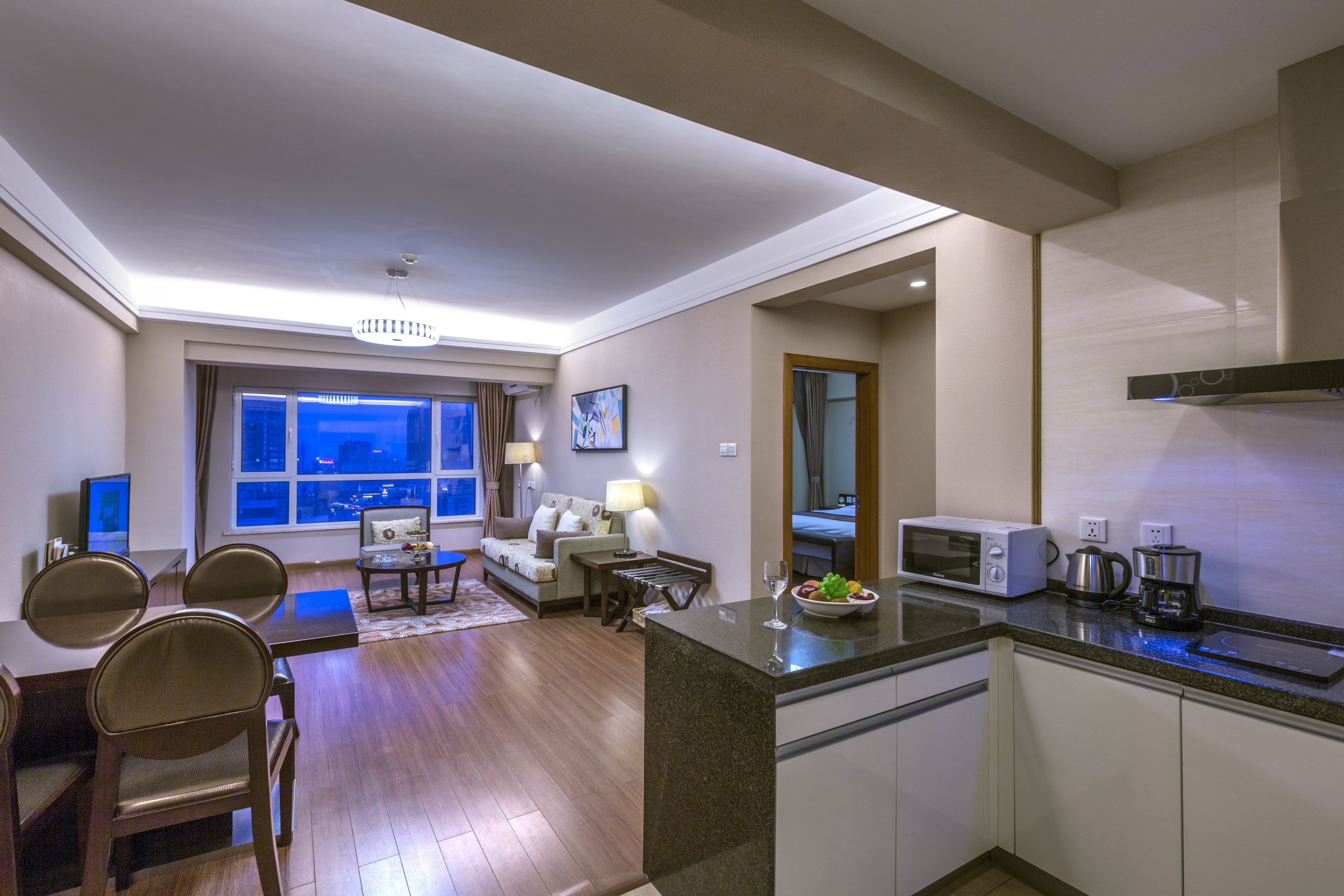 A modern hotel room with a dining area, kitchen, and a large window offering a city view.