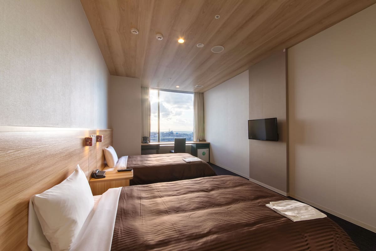 A hotel room with two beds, a TV, a desk with a lamp, and a window with a city view.