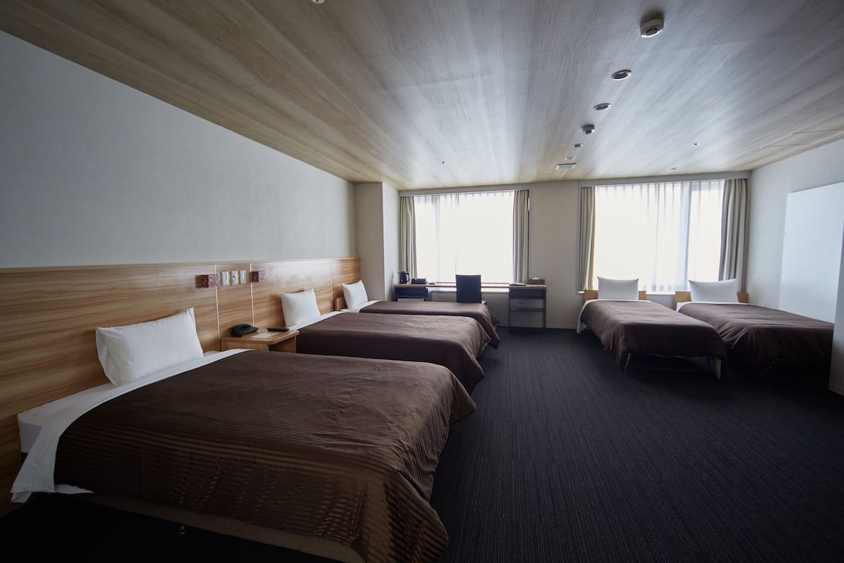 A hotel room with four beds, a desk, a chair, and a window with curtains.