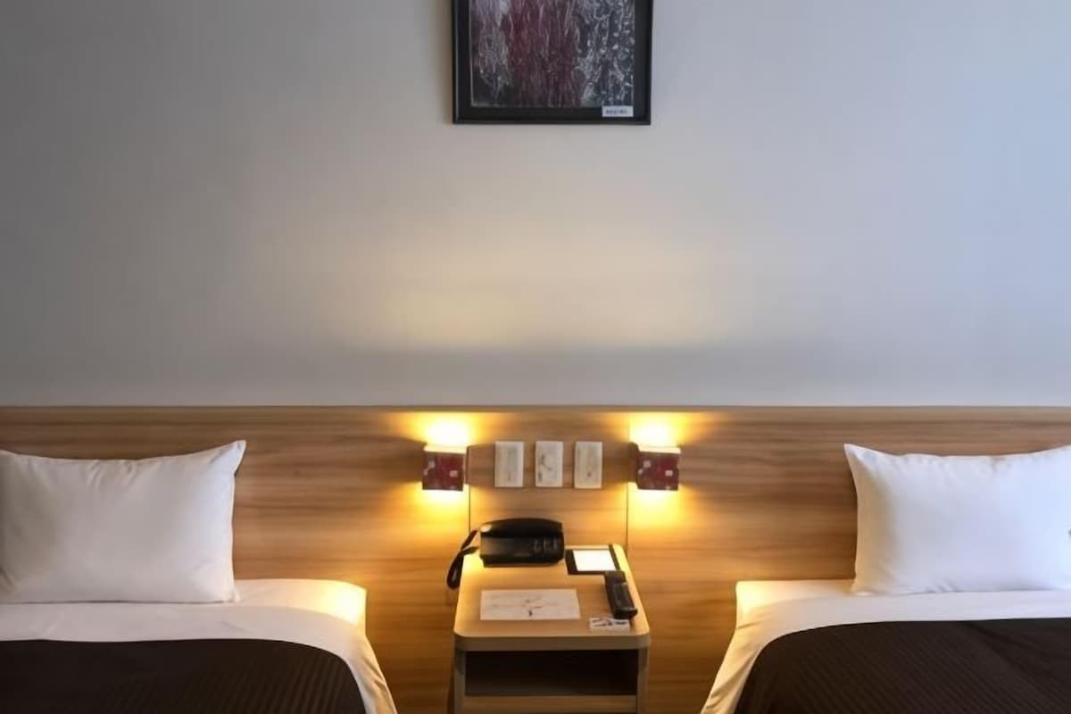 A hotel room with two beds, a bedside table with a lamp, and a framed picture on the wall.