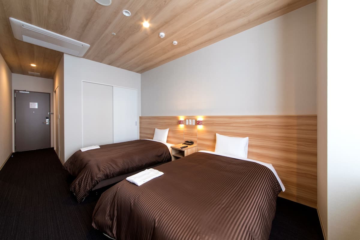 A hotel room with two beds, a wooden ceiling, a wall-mounted light, and a door.