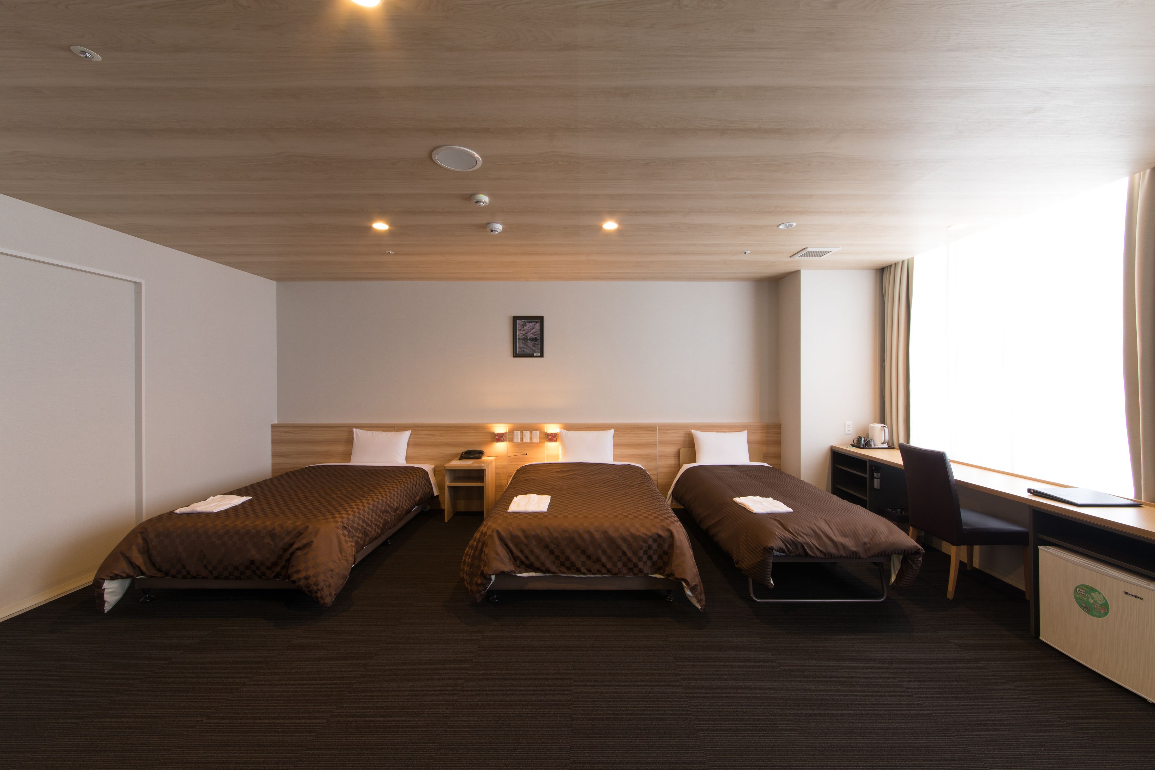 A hotel room with three single beds, a desk with a chair, a small table, and a window with curtains.