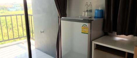 A compact room with a balcony, a small refrigerator, a shelf with bottles, and a trash bin.