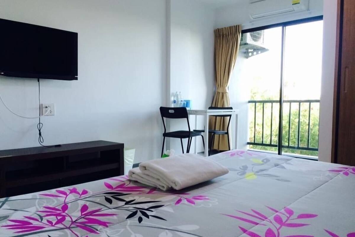 Habitación de hotel con cama, televisión montada en la pared, escritorio con silla y balcón con vistas a zonas verdes.