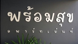 A wall with white letters spelling out a name in a non-Latin script, possibly Thai, with a plant in the foreground. - Promsook Apartment (Chonburi)