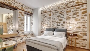 A bedroom with a stone wall, a bed with grey bedding, a bedside table, and a mirror. - 1844 Suites Syros (Syros)