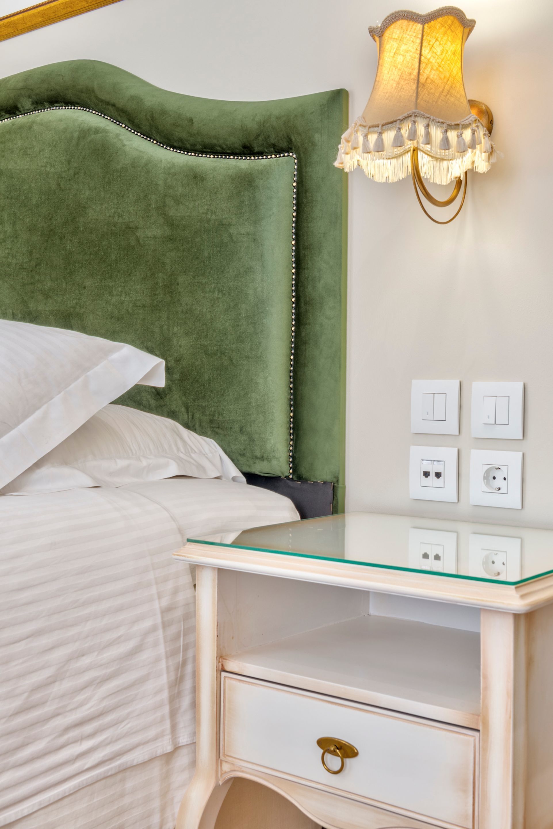 A bed with a green upholstered headboard, a bedside table with a glass top, and a wall-mounted lamp with a fringed shade.