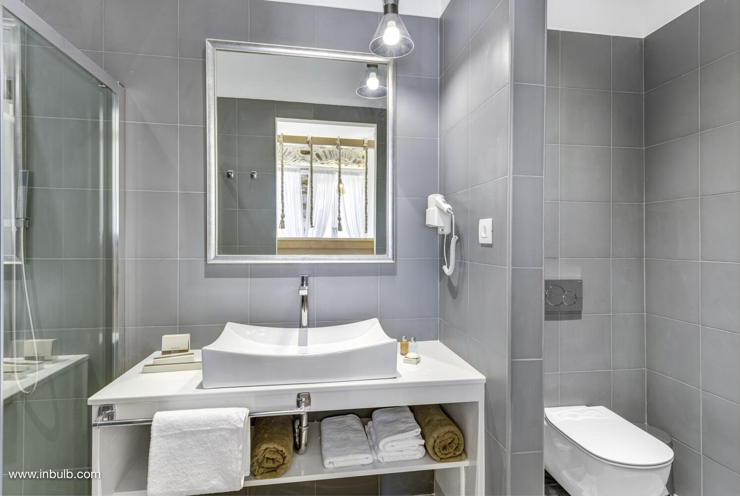 Un baño moderno con un lavamanos blanco, espejo y retrete. Hay un perchero con toallas y una mampara de cristal para la ducha.