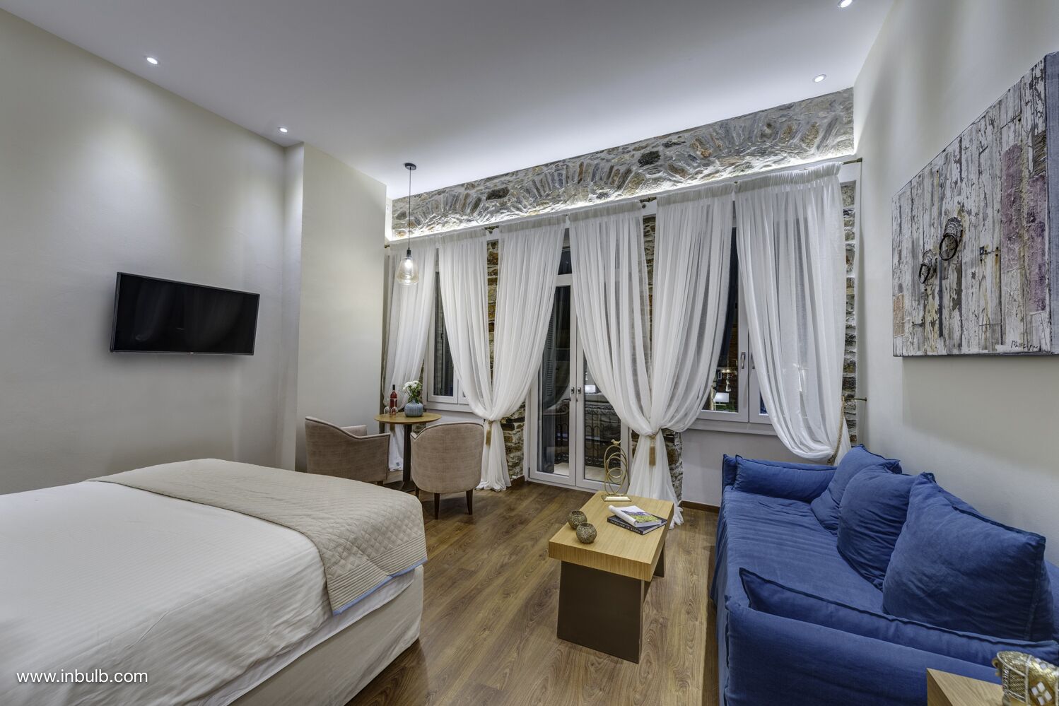 A modern hotel room with a bed, a blue sofa, a small table, a TV, and a window with curtains.