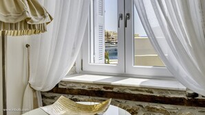 A room with a view of a marina and buildings, featuring a window with white curtains, a wooden bench, and a white table with a woven basket. - 1844 Suites Syros (Syros)