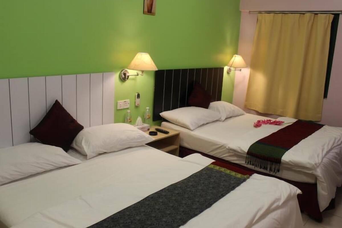 A hotel room with two beds, each with white linens and a dark-colored runner. There are two bedside lamps, a nightstand with a bottle and tissues, and a window with yellow curtains.