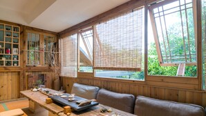 Ein traditioneller japanischer Raum mit Tatami-Bodenbelag, einem niedrigen Holztisch mit Teezeremonie-Gefäßen, einer gepolsterten Sitzgelegenheit und großen Fenstern mit Bambusrollos.
