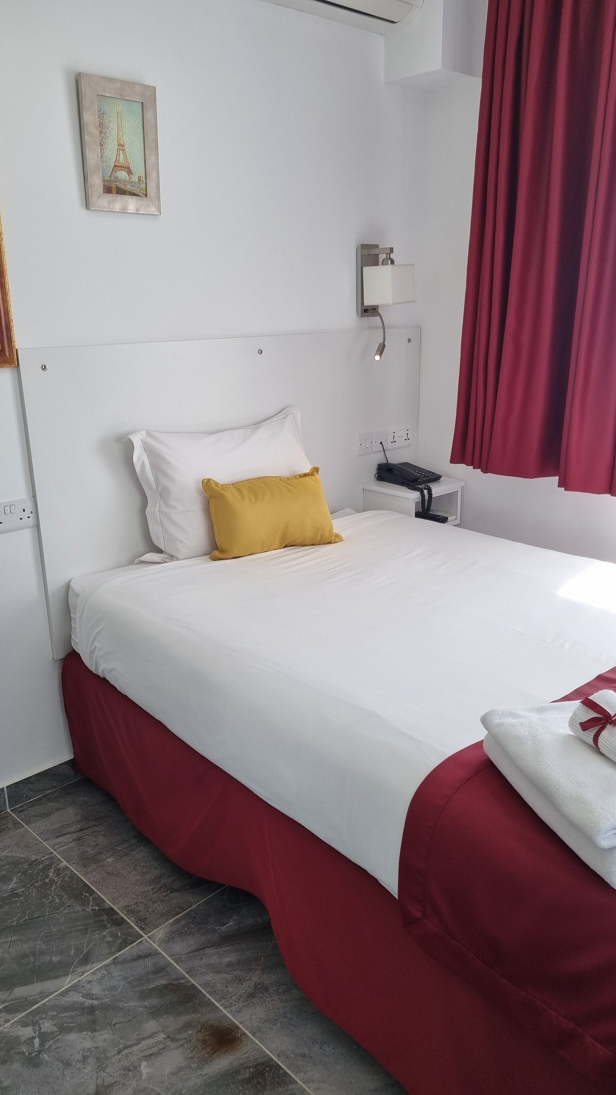 A hotel room with a bed, a nightstand, a wall-mounted lamp, a power outlet, and a red curtain.