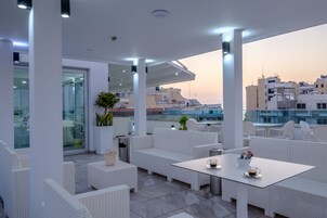 Un patio en la azotea con mobiliario de mimbre blanco, una entrada con puerta de vidrio y vistas a la ciudad.