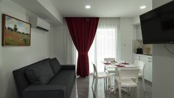 Un salon moderne avec un canapé gris foncé, un coin repas avec une table et des chaises blanches, un rideau rouge et un téléviseur fixé au mur.