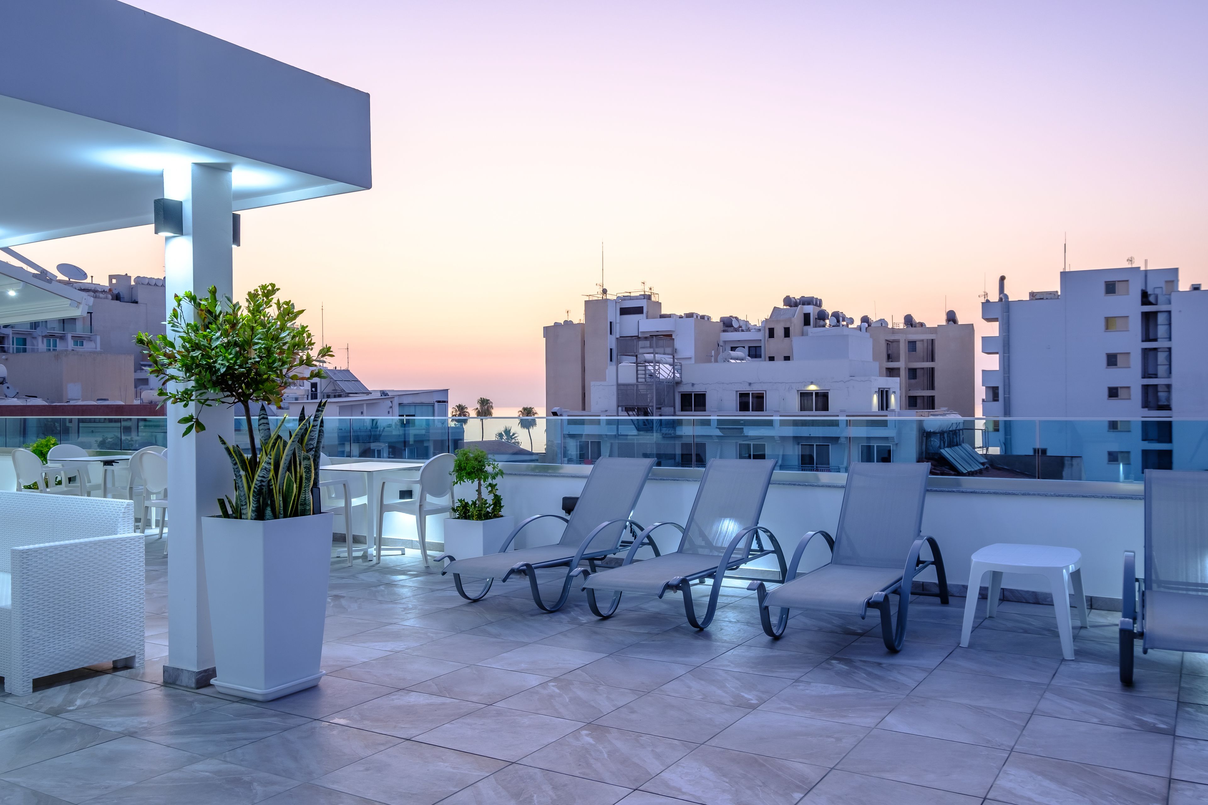 A rooftop terrace with lounge chairs and potted plants, overlooking a cityscape at sunset.