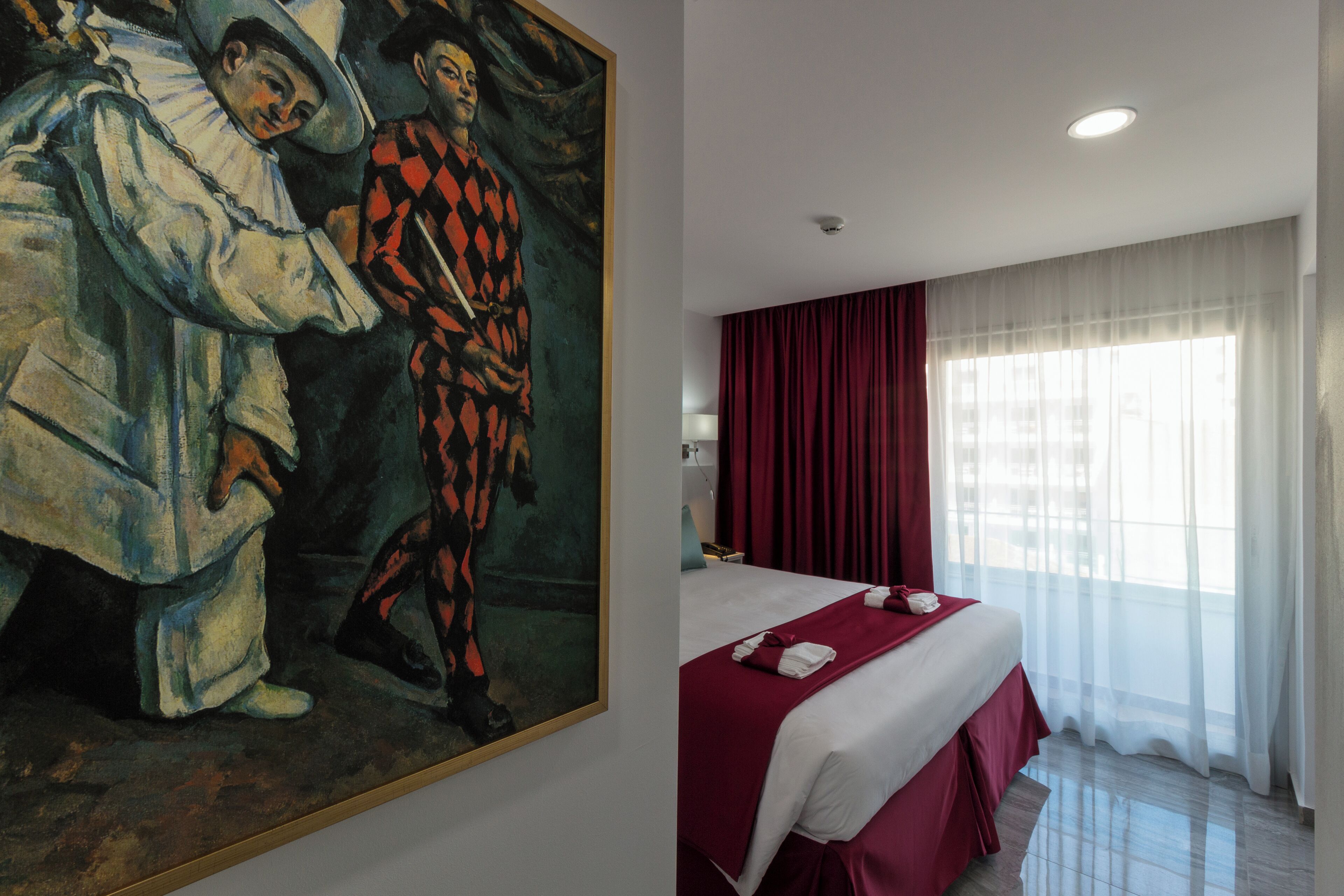 A hotel room with a large painting on the wall, a bed with a red bedspread, and a window with sheer curtains.