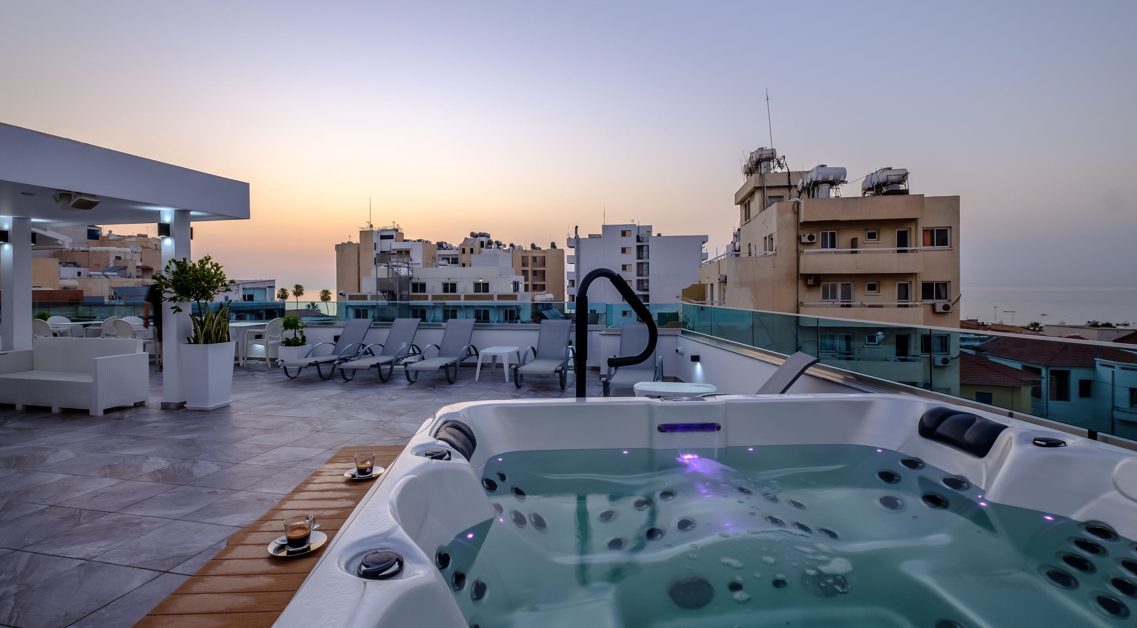 A rooftop terrace with a hot tub, lounge chairs, and a view of the cityscape at sunset.