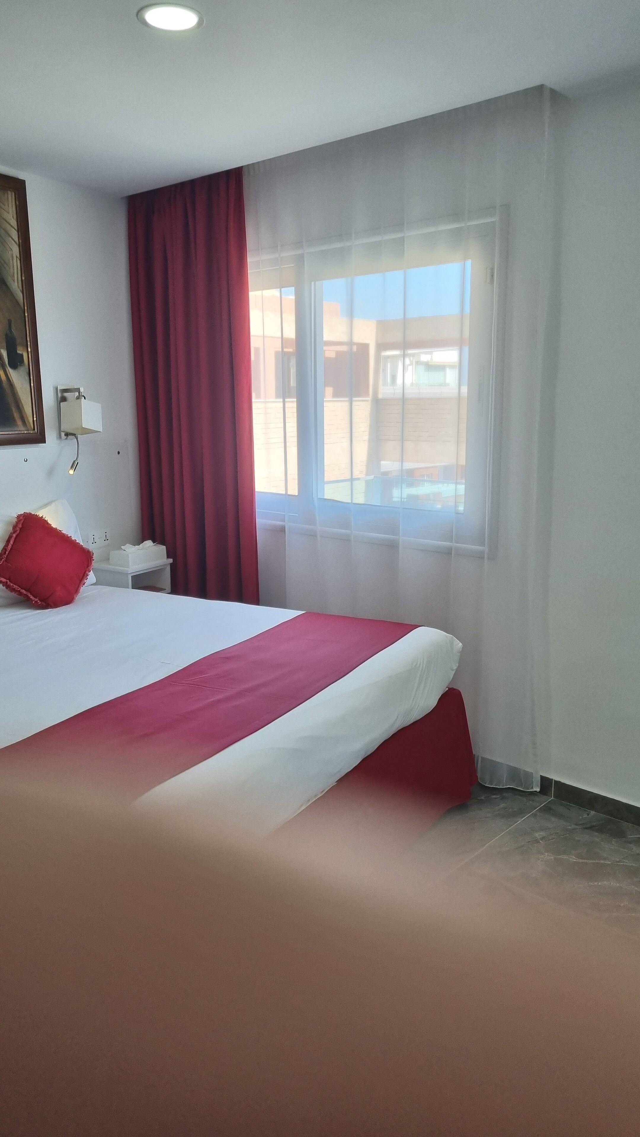 Habitación de hotel con una cama grande, una mesita de noche, una lámpara, una ventana con cortinas y un cuadro en la pared.