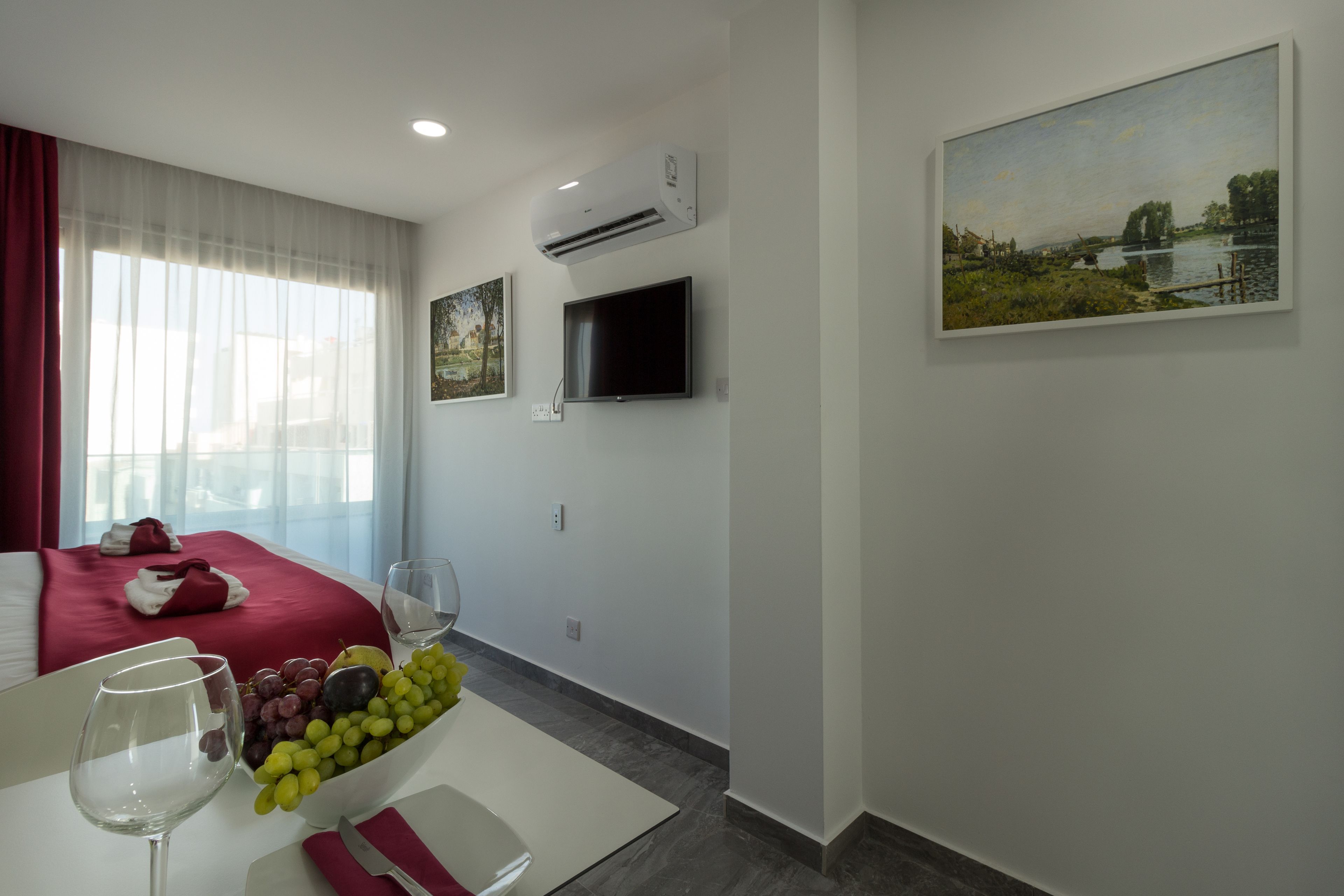 A hotel room with a bed, a flat-screen TV, an air conditioning unit, a painting on the wall, and a fruit bowl on the table.