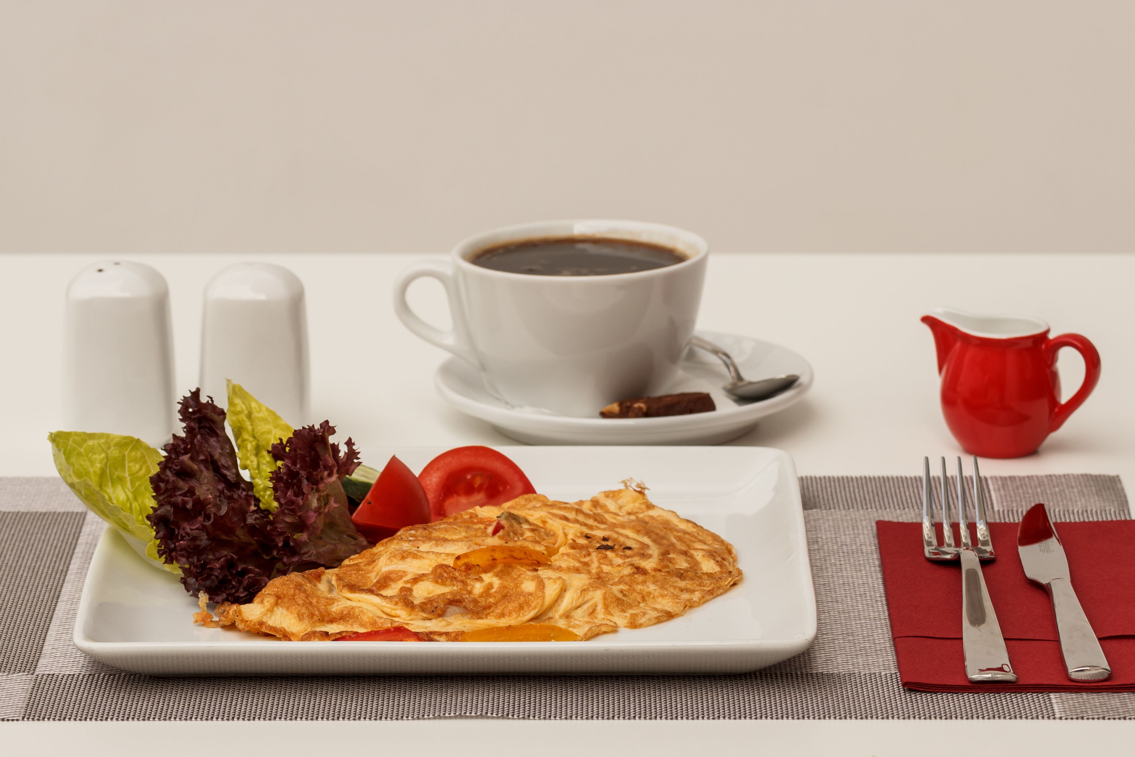 A breakfast setting with a plate of food, a cup of coffee, and a red pitcher.