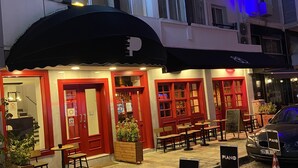 Exterior de un hotel con fachada roja y blanca, mobiliario exterior y un cartel de piano.