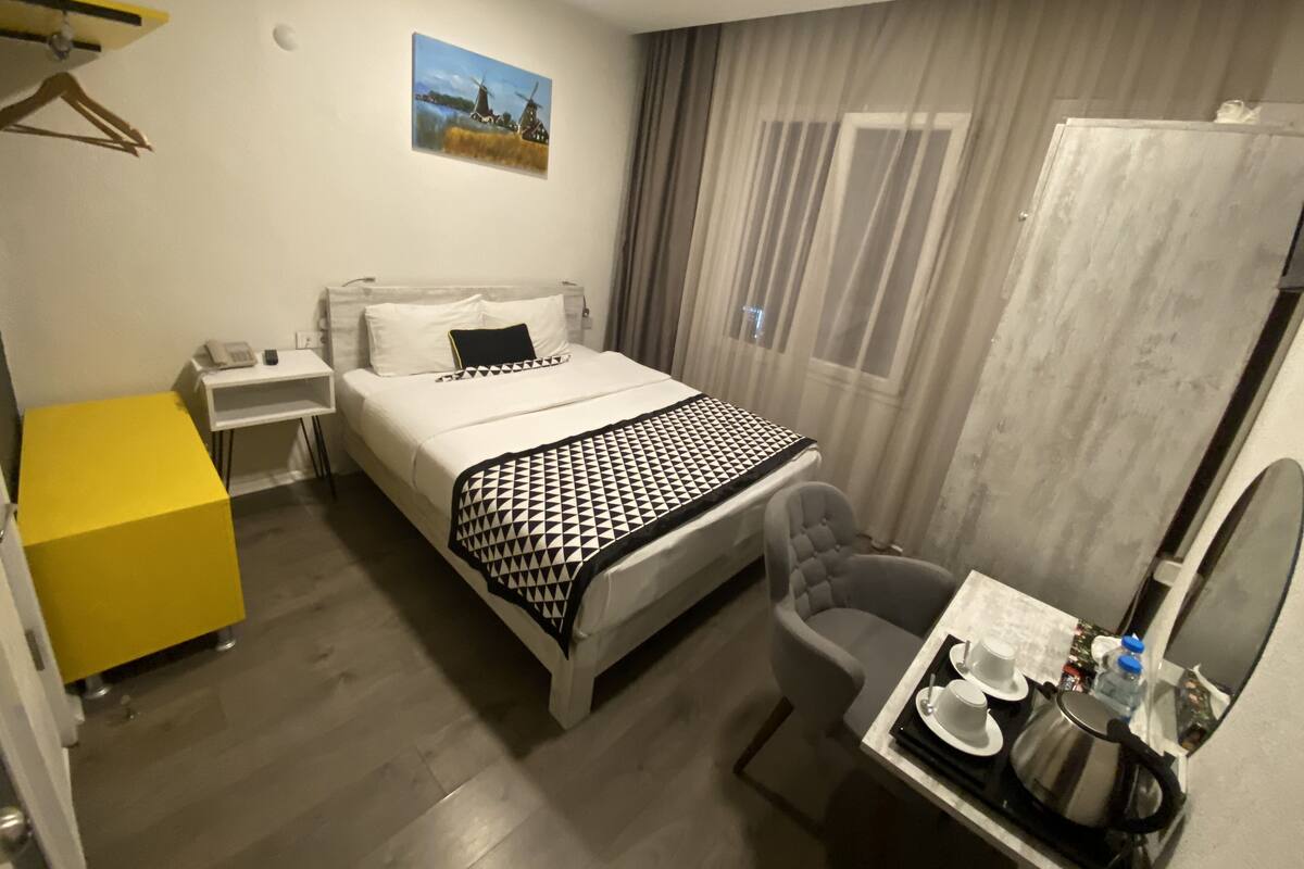 A hotel room with a bed, bedside table, chair, and a small table with a kettle and cups.