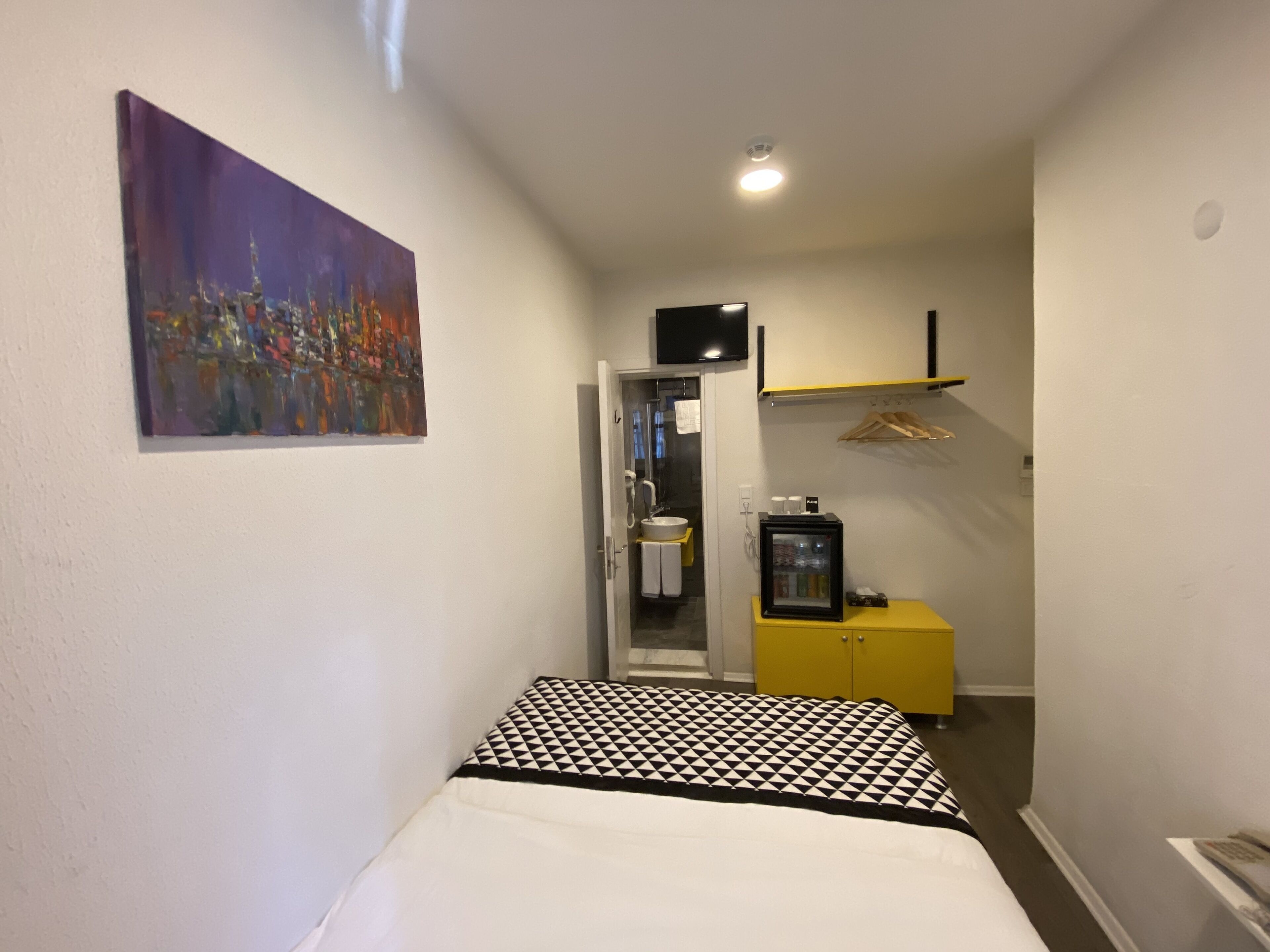 Ett litet, smalt hotellrum med en säng, en tavla på väggen, ett badrum med handfat och spegel samt ett litet gult skåp med ett kylskåp.