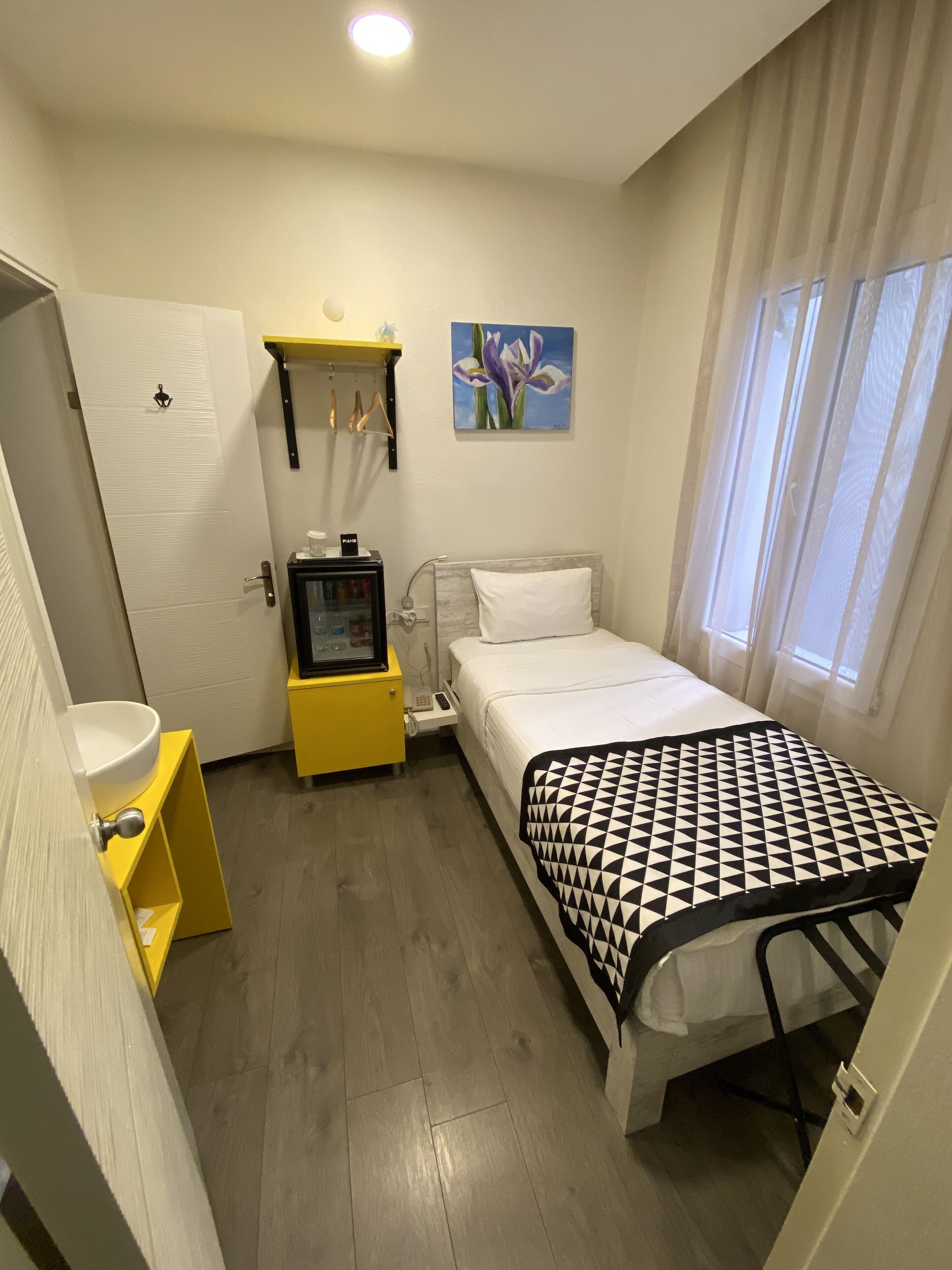 Habitación de hotel compacta con cama, mesita de noche y un escritorio pequeño con silla. Hay una ventana con cortinas, un cuadro en la pared y una puerta con manija.