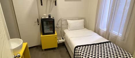 Habitación de hotel compacta con cama, mesita de noche y un escritorio pequeño con silla. Hay una ventana con cortinas, un cuadro en la pared y una puerta con manija.