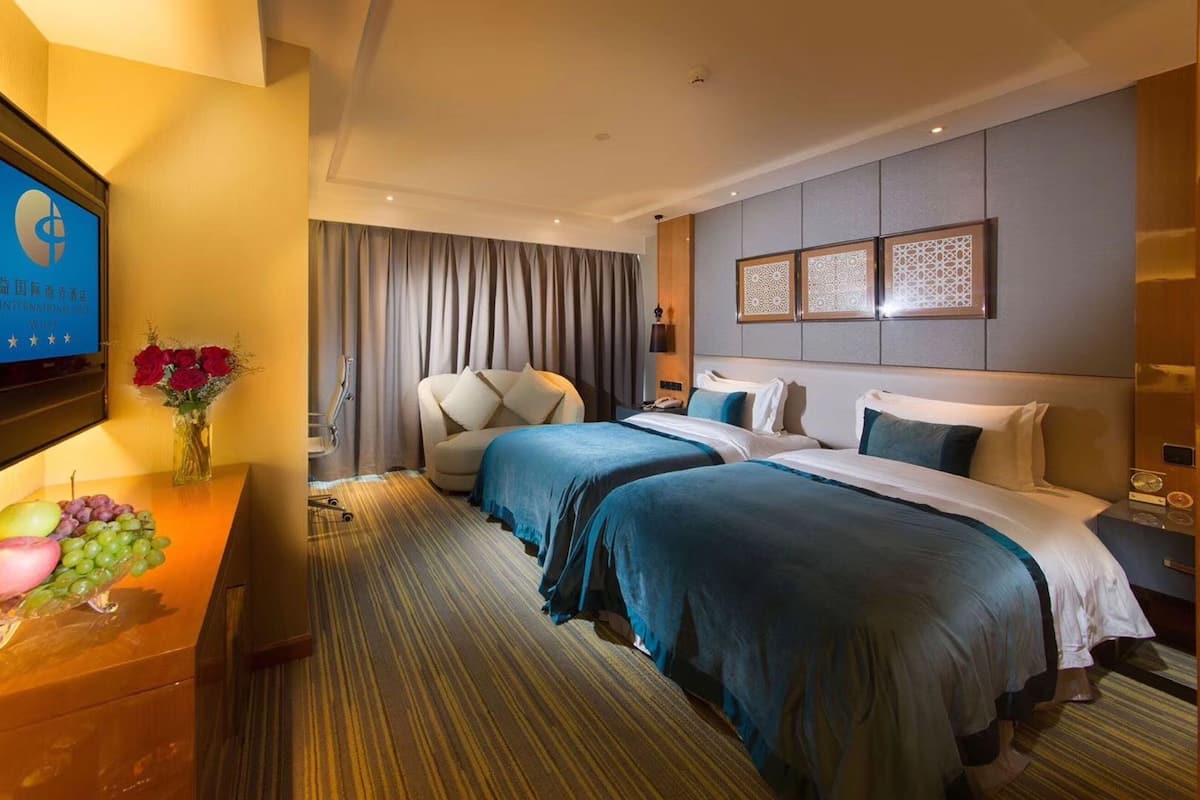 A hotel room with two beds, a desk with a TV, a vase of flowers, and a view of the city.