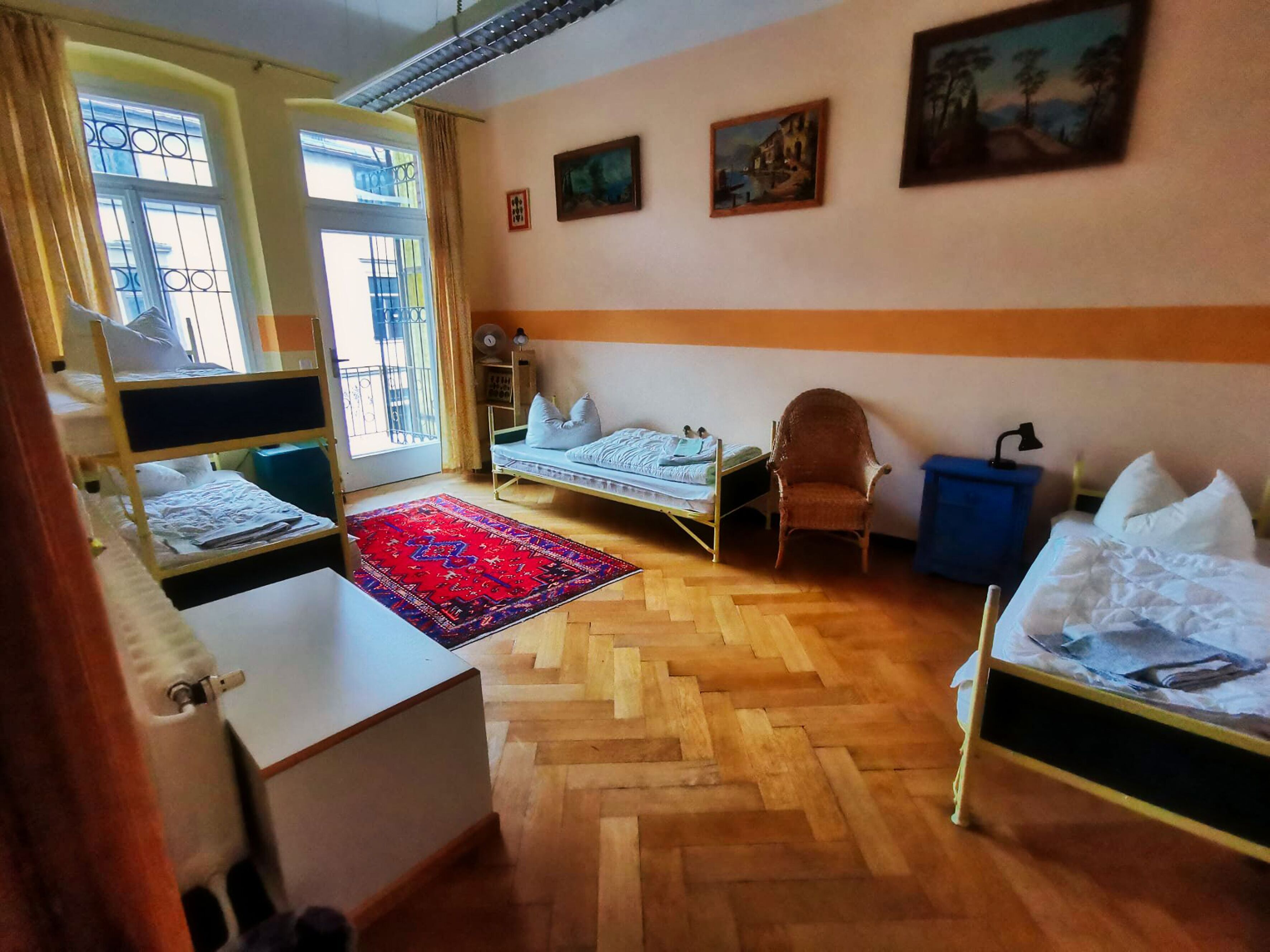Una habitación con literas, un escritorio, una silla, una mesita y un tapete rojo, además de una ventana con cortinas.