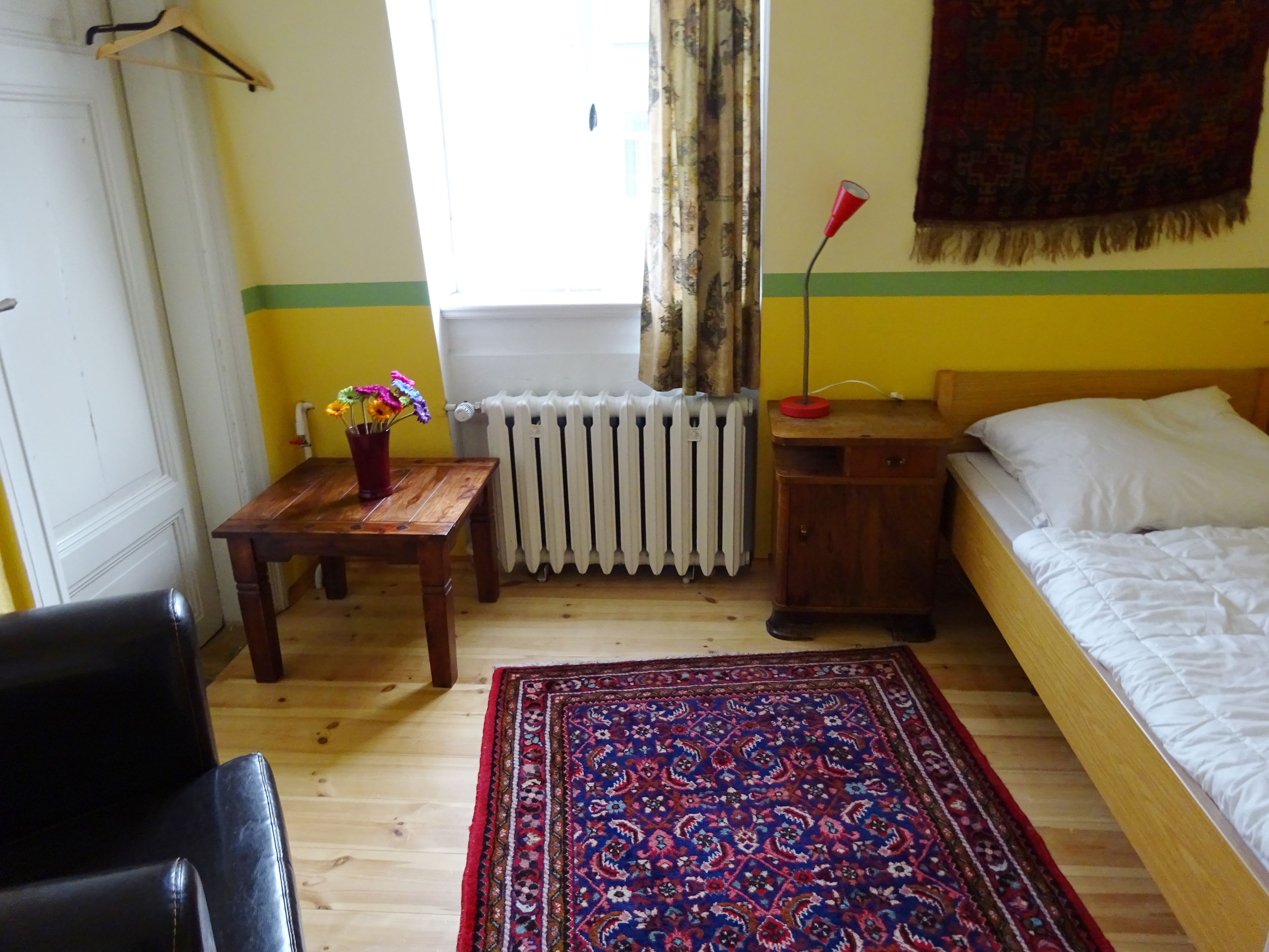 Ein Zimmer mit einem Bett, einem Holztisch mit Blumenvase, einem Heizkörper, einer Hängelampe und einem gemusterten Teppich.