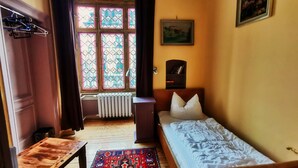 A room with a bed, a wooden table, a radiator, a window with curtains, a bookshelf, and a painting on the wall. - Hummel Hostel (Weimar)