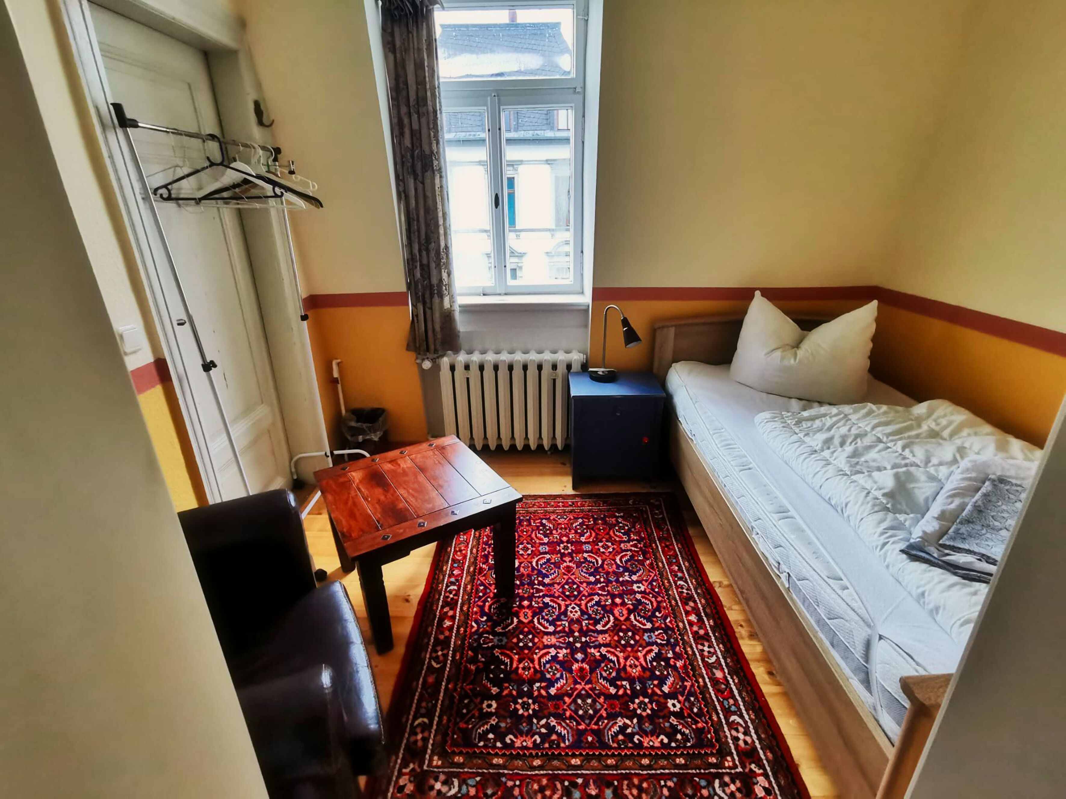 Ein kleiner, gemütlicher Raum mit Bett, Tisch, Stuhl, Heizkörper und einem fenster mit Vorhängen.