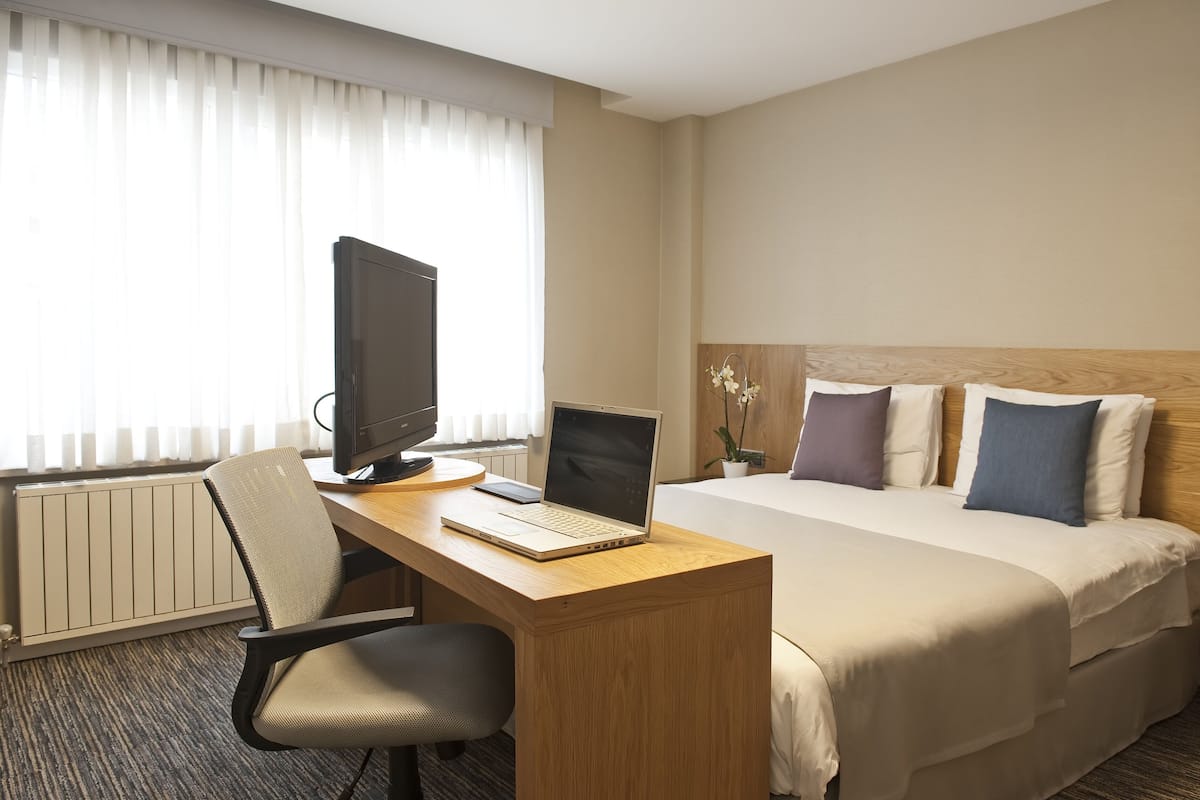 Et hotelværelse med en seng, et skrivebord med computer, en stol og et vindue med persienner.
