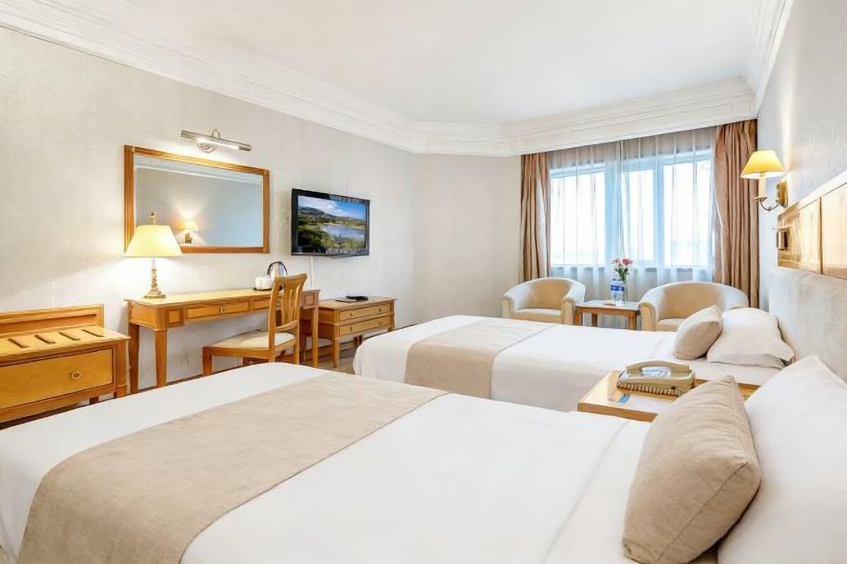 A hotel room with two beds, a desk, a chair, a TV, and a window with curtains.