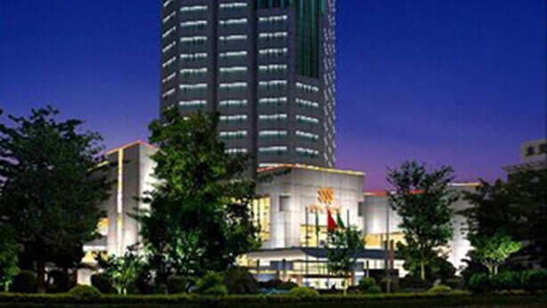 A tall, modern building with a distinctive spire at the top, surrounded by greenery and other buildings. - Hotel Silverland (Dongguan)