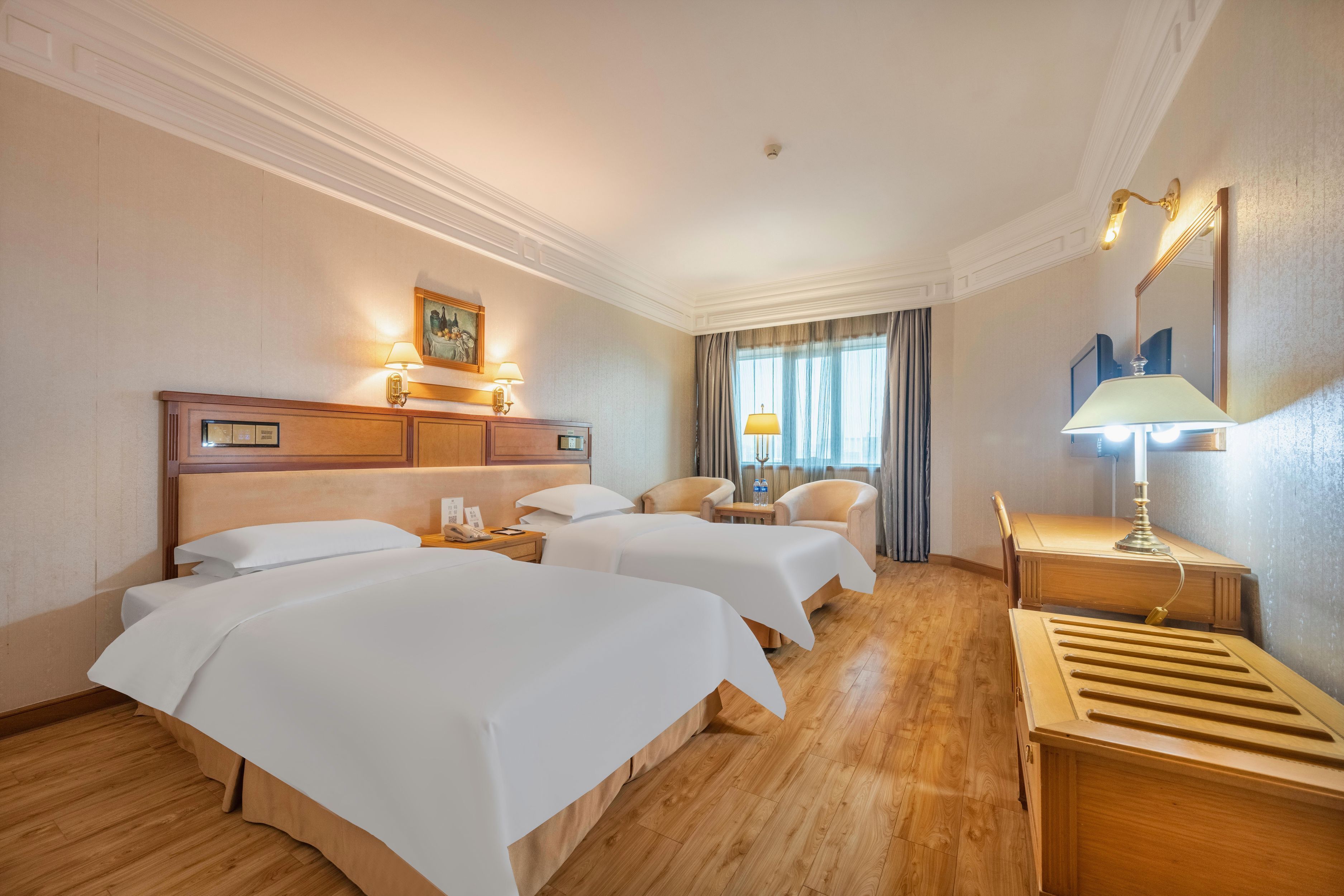 A hotel room with two beds, a desk, a chair, and a lamp.