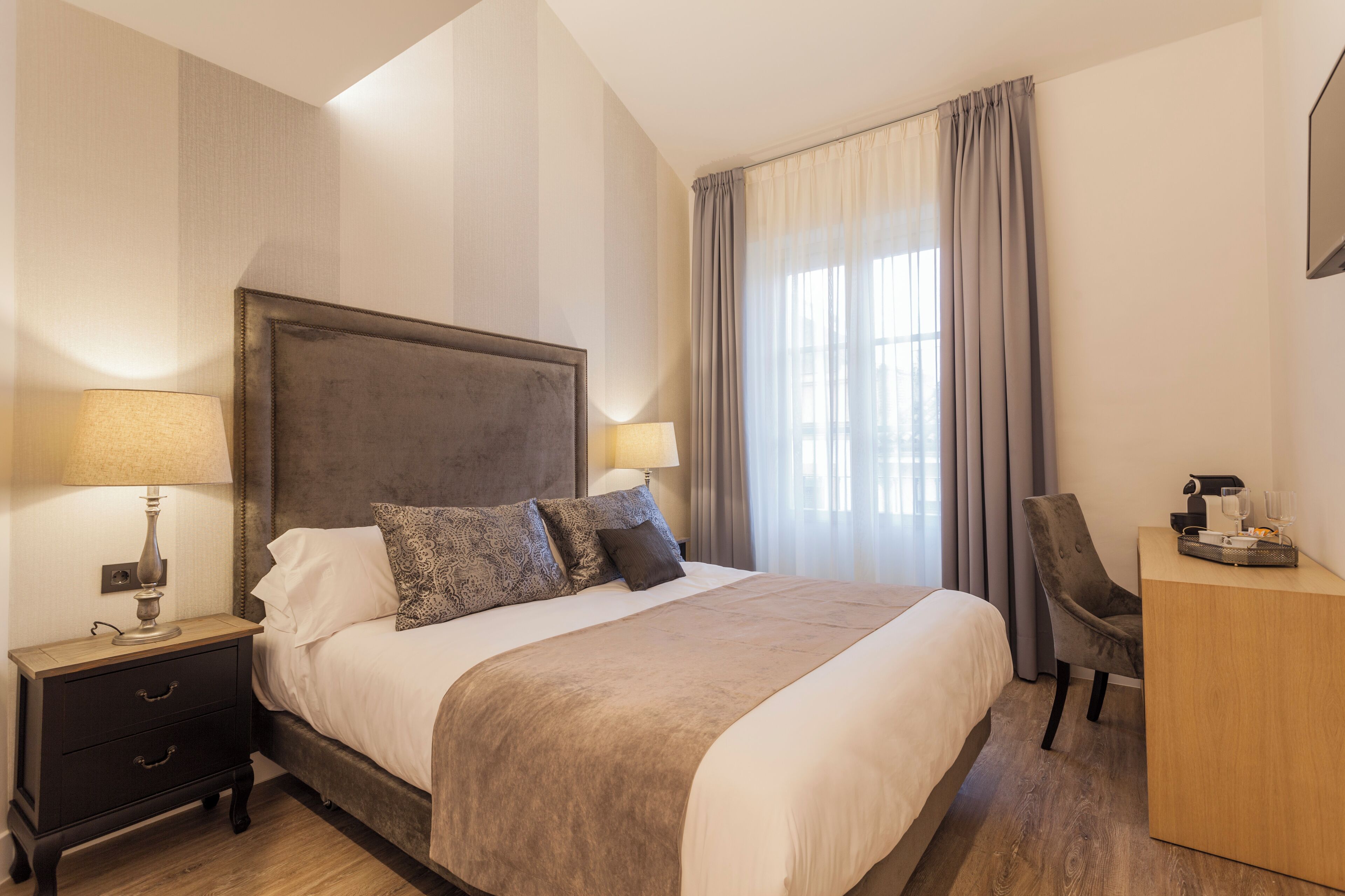 A neatly made bed with a headboard, a bedside table with a lamp, a desk with a chair, and a large window with curtains.