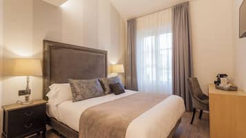A neatly made bed with a headboard, a bedside table with a lamp, a desk with a chair, and a large window with curtains.