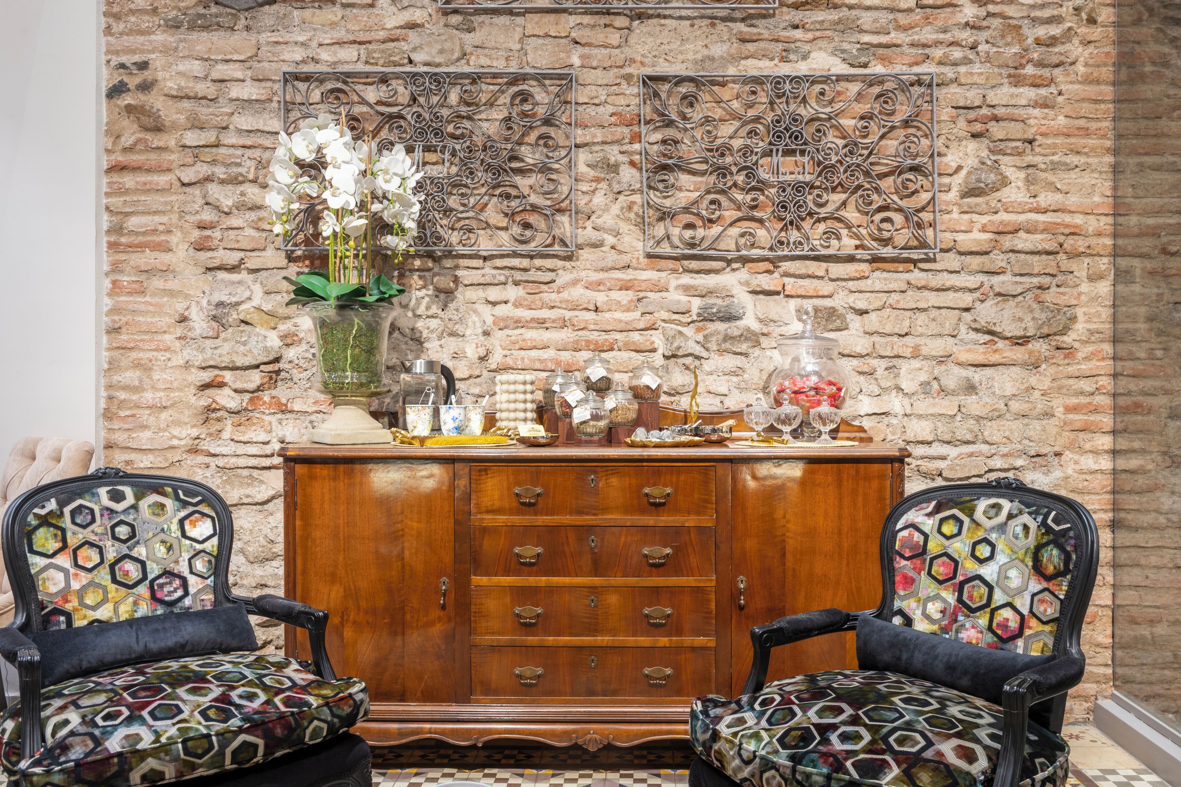 A room with two patterned armchairs, a wooden cabinet with drawers, and decorative wall art against a brick wall.