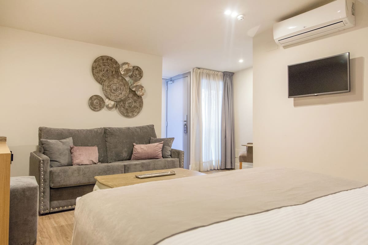 Une chambre d’hôtel moderne avec un canapé gris, un téléviseur à écran plat, un appareil de climatisation et un lit.