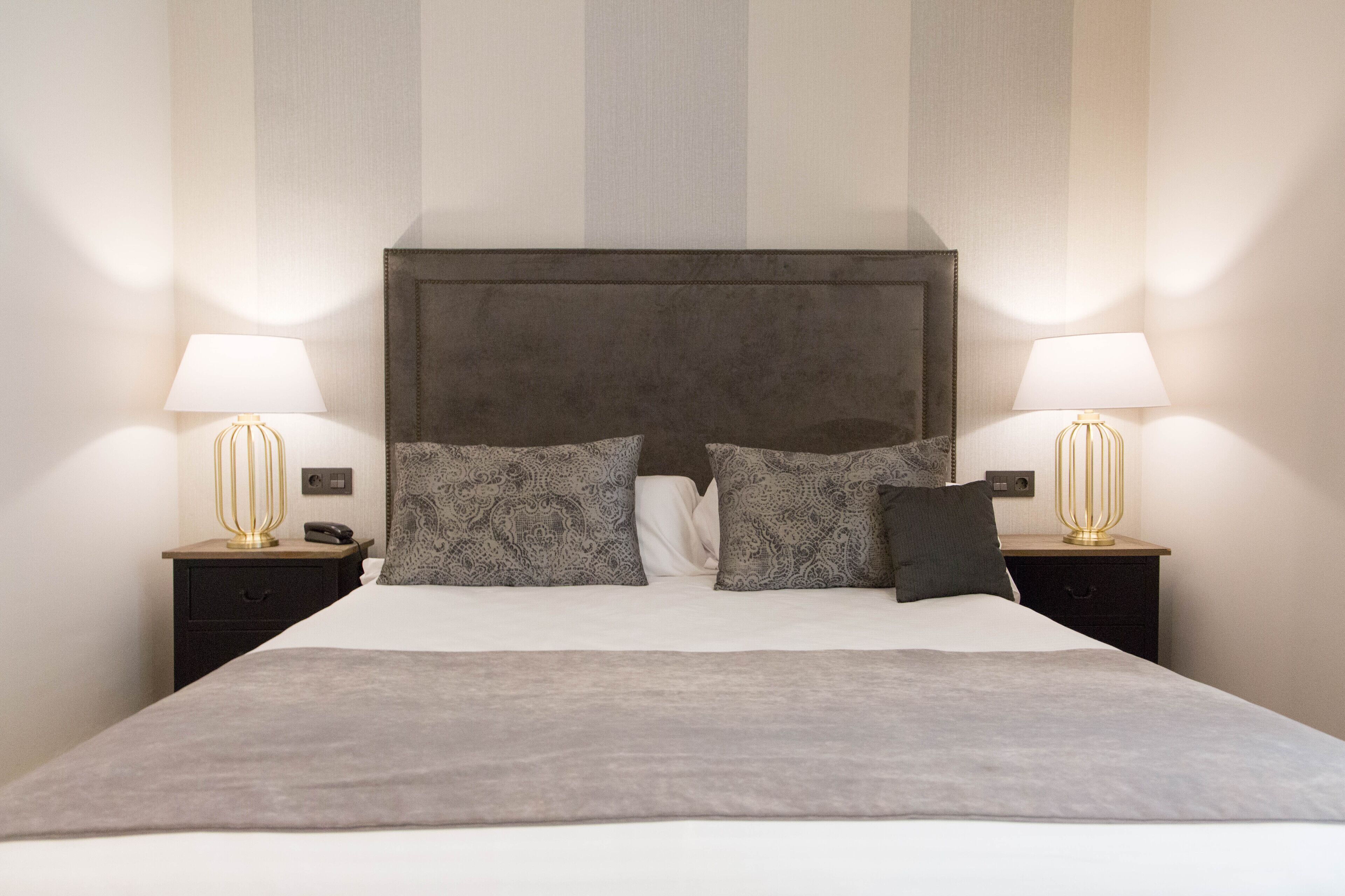 A neatly made bed with a grey blanket, two patterned pillows, and a dark headboard. There are two bedside tables with lamps and a telephone on each. The walls have a striped pattern.