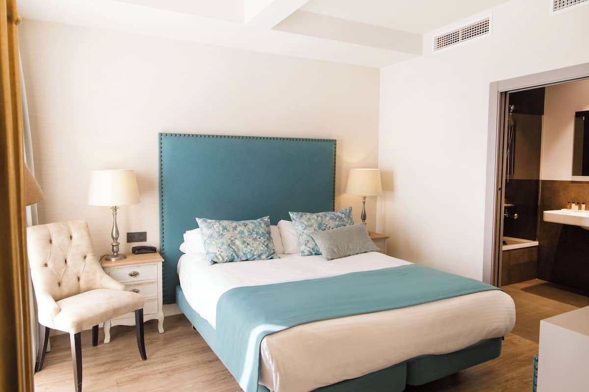 Une chambre d’hôtel avec un grand lit, deux tables de chevet avec des lampes, une chaise et une salle de bain visible à travers une porte ouverte.
