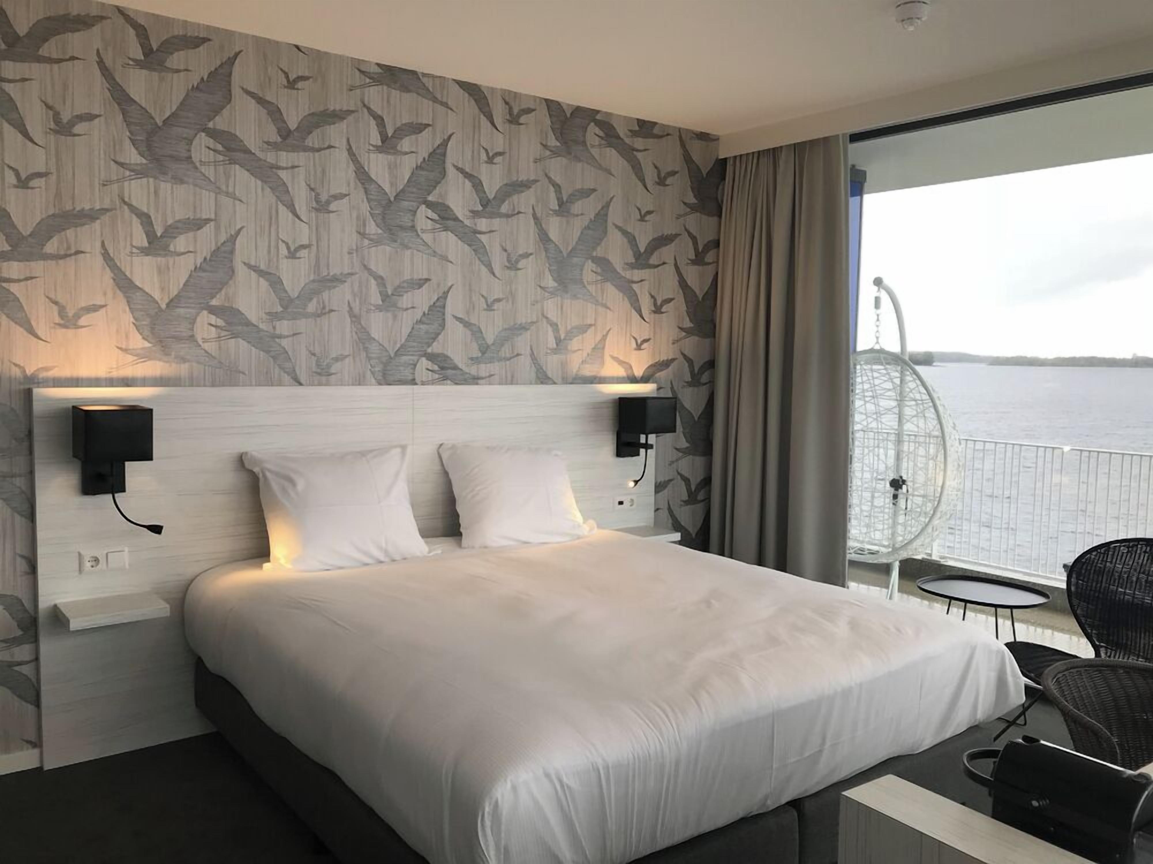 Une chambre d’hôtel avec un grand lit, deux lampes fixées au mur, une vue sur l’eau et un balcon avec une chaise.