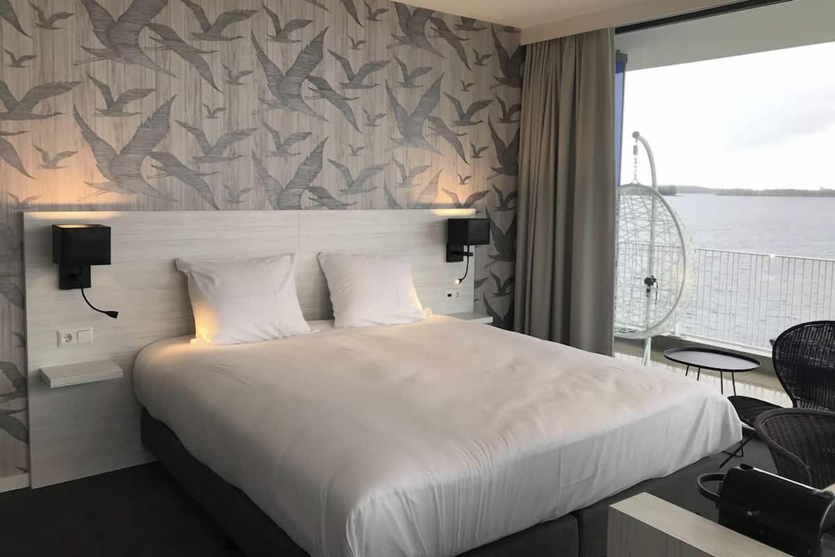 Ein Hotelzimmer mit einem großen Bett, zwei Wandleuchten, Blick auf das Wasser und einem Balkon mit einem Stuhl.