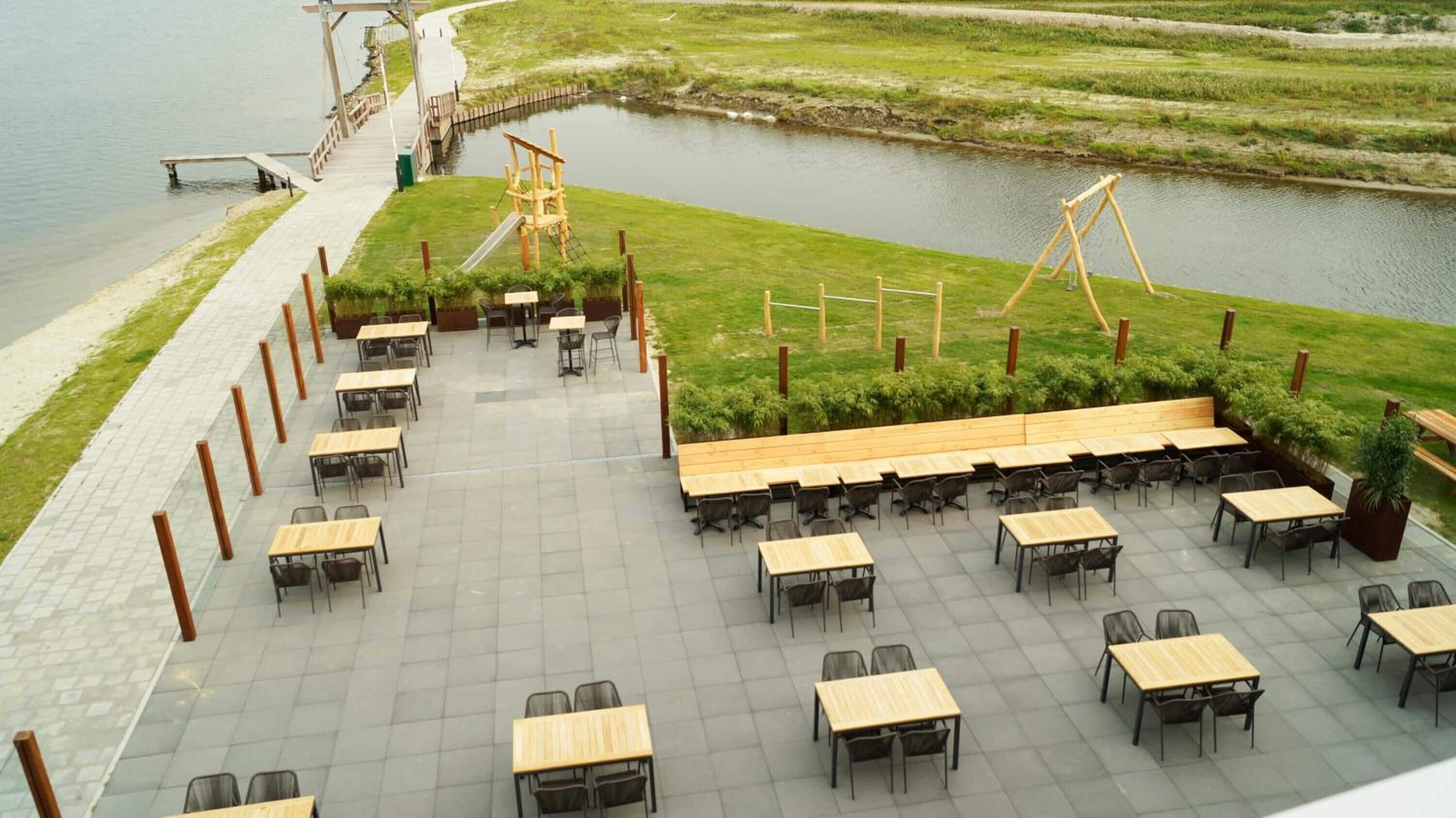 An outdoor dining area with tables and chairs, a wooden swing set, and a view of a water body.